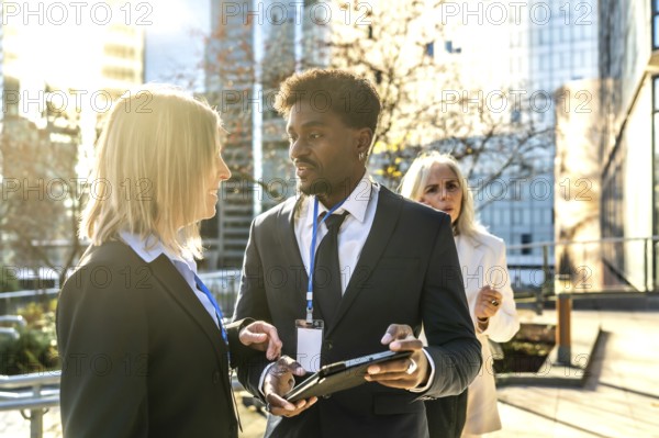 Diverse business professionals networking and collaborating while reviewing information on a digital tablet outdoors in an urban setting, with a female colleague walking behind them