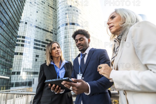 Diverse business professionals collaborating on a digital tablet, standing outdoors in front of modern glass skyscrapers, symbolizing city life, teamwork, and global business strategies
