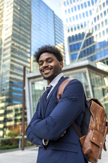 Young black businessman standing confidently in a modern urban environment, carrying a brown leather backpack and smiling brightly, portraying success, ambition, and professional growth