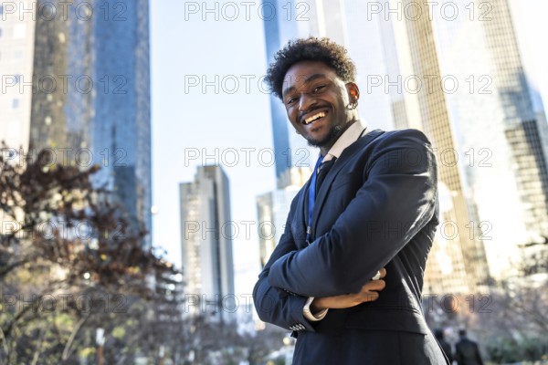 Young adult african american businessman standing with arms crossed. Smiling broadly at the camera. Projecting confidence and success against a backdrop of modern skyscrapers in a sunny urban setting