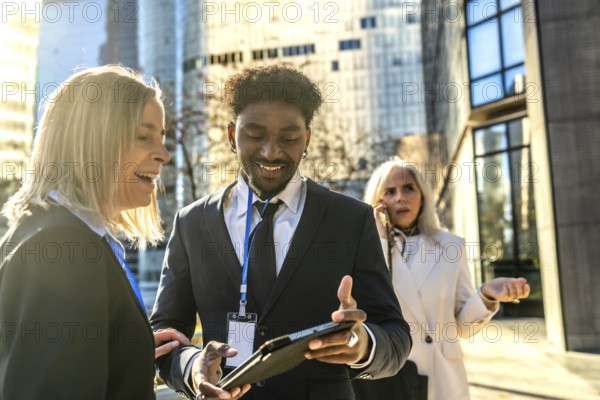 Diverse business people working together outdoors in a city, exchanging ideas and smiling while looking at a tablet, with another colleague talking on the phone in the background