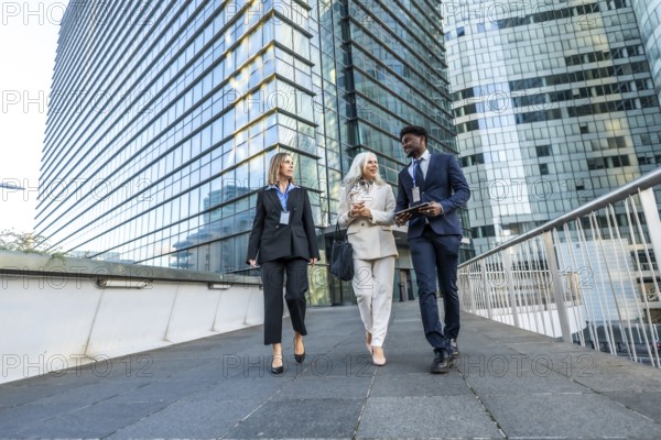 Three multi ethnic business colleagues walking and talking on a pedestrian bridge amid modern glass offices, showcasing urban collaboration, networking and professional teamwork
