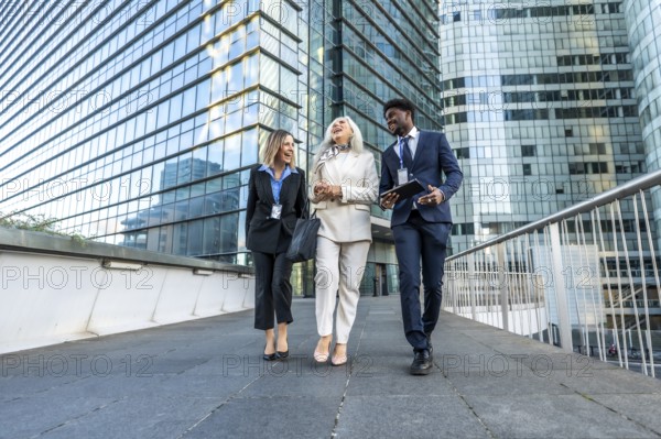Three diverse business professionals. Including a young black man. A mature woman. And a young woman. Are walking and laughing outdoors in a modern city's financial district. Collaborating on projects