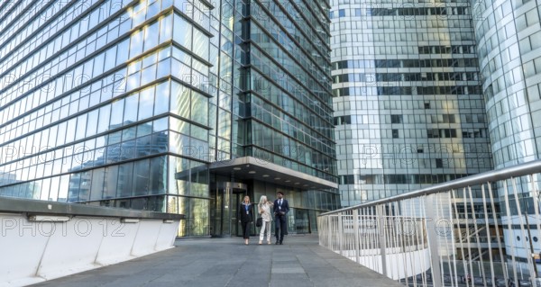 Business professionals walking together on an elevated pedestrian walkway, surrounded by contemporary glass skyscrapers and corporate headquarters, representing teamwork and urban success