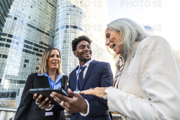 Diverse business professionals smiling and interacting, using a digital tablet for outdoor collaboration and teamwork in a modern city setting with skyscrapers