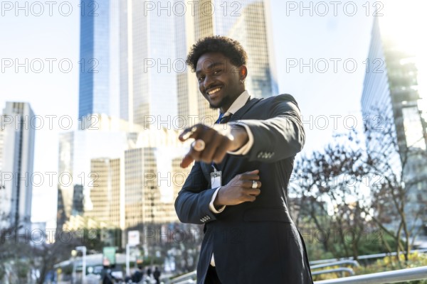 Young black businessman smiling and pointing directly towards the viewer with one hand, standing outdoors in a modern city setting with skyscrapers in the background during a bright day