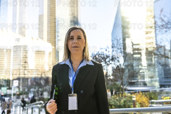 Professional businesswoman standing outdoors in a modern downtown area, wearing a suit and lanyard identification badge, representing corporate success and career progress