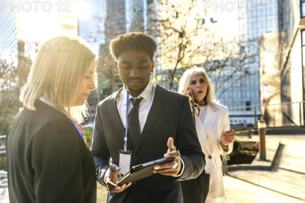 Business colleagues collaborating on an outdoor urban meeting, exchanging ideas while reviewing information on a digital tablet, with a female coworker talking on a mobile phone in the background
