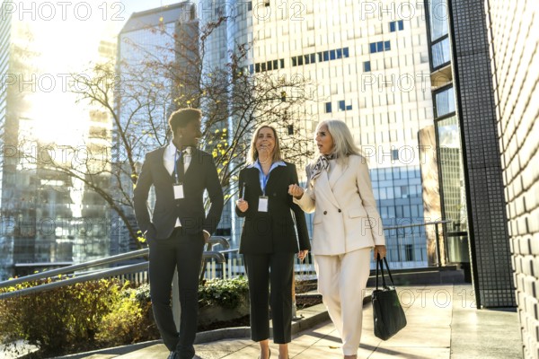 Diverse business professionals walking outdoors on a sunny day, engaging in conversation while heading to work in a modern corporate urban environment with skyscrapers