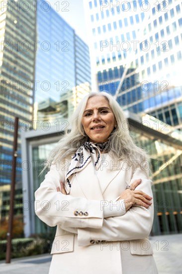 Senior businesswoman with gray hair in a cream suit stands confidently with arms crossed before modern office towers, projecting leadership, experience and urban professional success