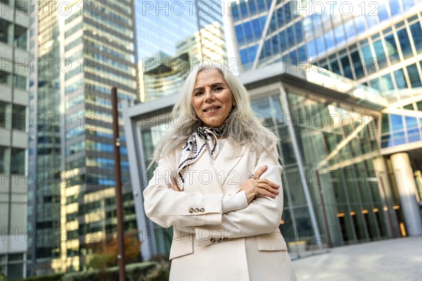 Senior businesswoman with gray hair and a formal elegant blazer standing outdoors with folded arms, portraying confidence and leadership in a vibrant metropolitan financial district