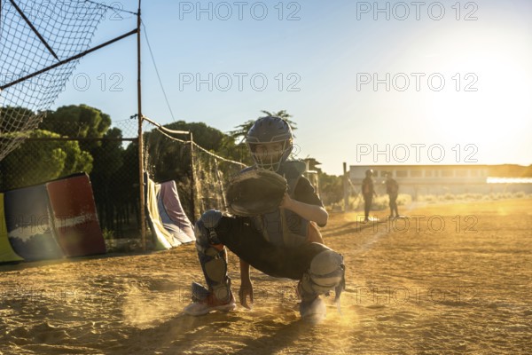 Boy catcher wearing protective gear and mitt, squatting on a dusty baseball field with golden backlighting, focused on the game during practice or youth sports