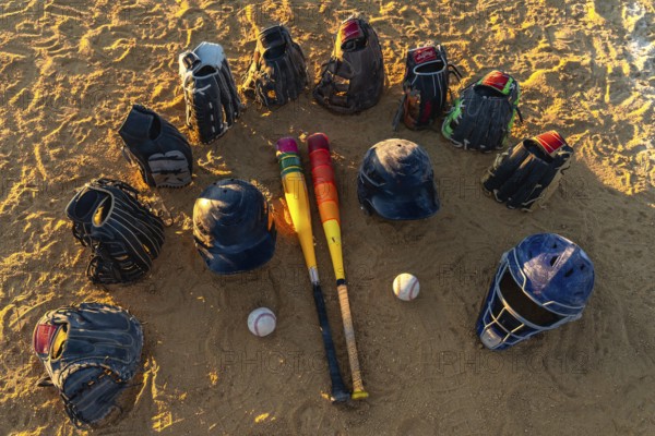 Baseball bats, gloves, helmets and catcher's mask arranged in a circle on a sandy field at sunset, warm light highlighting gear ready for team practice and play