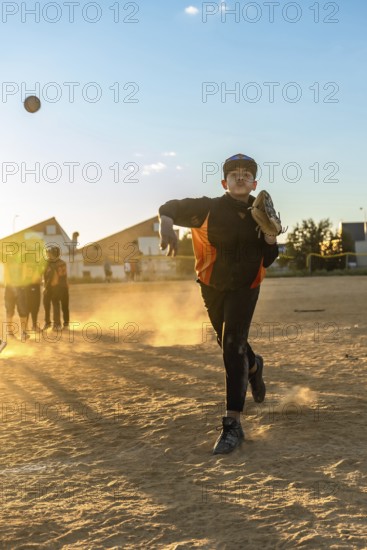 Young boy in baseball uniform pitching a baseball, creating a cloud of dust on the field during an evening game with other players in the blurred background