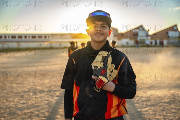 Young male baseball player stands confidently on a sunlit dirt field at golden hour, cap on and glove in hand, smiling and ready for practice or a neighborhood game