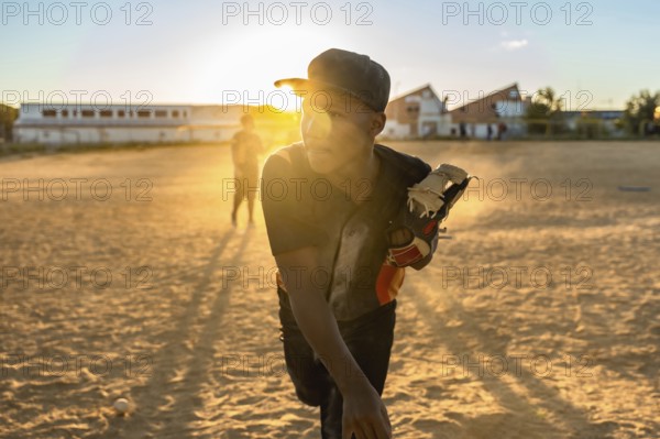 Young baseball player focusing while preparing to throw from the pitcher's mound, wearing a cap and glove, with golden light from the setting sun casting long shadows on the dirt field
