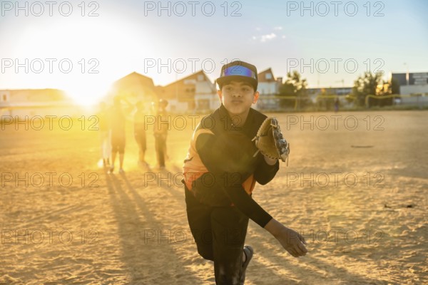 Young boy in baseball uniform and glove pitching a baseball on a dirt field during sunset, with other players blurred in the background and warm lens flare