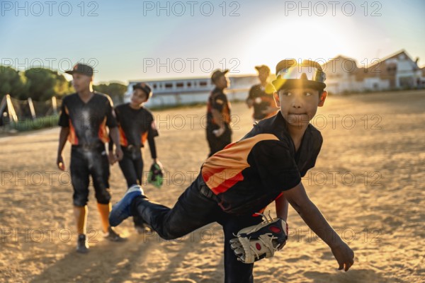 Young baseball player pitching the ball on a dusty field with teammates standing by, wearing a cap and sports goggles, enjoying the game during a beautiful golden hour sunset