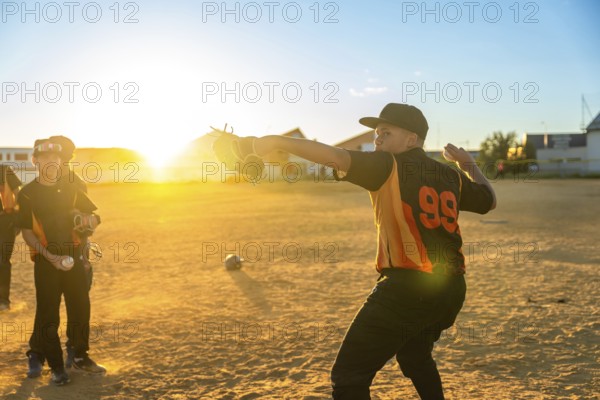 Young baseball player throwing a pitch with a glove during practice, with other players waiting on a dusty field under the warm glow of the setting sun