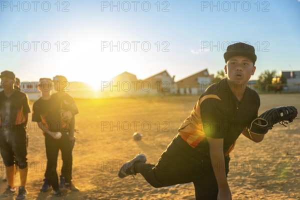 Young baseball pitcher in uniform and cap winds up to throw on a dusty field at golden sunset, teammates blurred behind him practicing and preparing in warm evening light