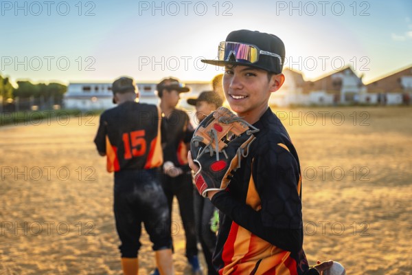 Young baseball player standing on a dirt field, holding a glove, smiling at the camera during a golden hour sunset with teammatesblurred in the background, representing youth sports and teamwork
