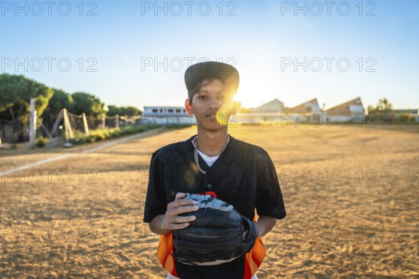 Young baseball player stands on a dusty field at golden hour, wearing a black uniform and cap, holding his glove, focused and ready to practice or play as the sun sets