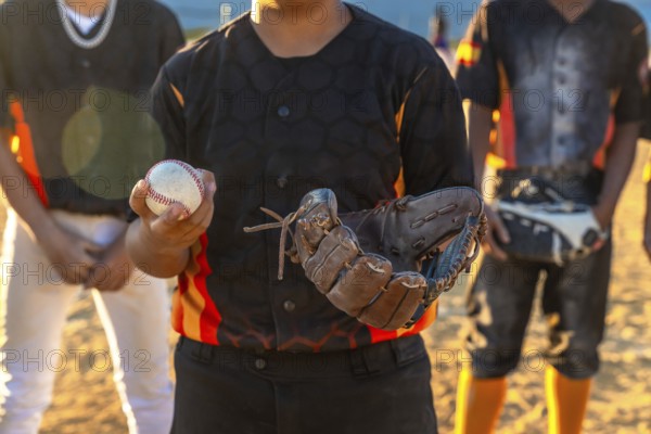 Young baseball player standing on a dusty field, holding a baseball in one hand and a catcher's mitt in the other, with blurred teammates in the background, ready for game or practice