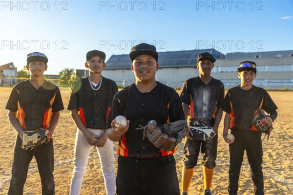 Group of diverse teenage baseball players standing on a dusty field, one holding a ball and glove, preparing for practice or a game during golden hour