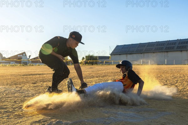 Two young baseball players engaged in a competitive game, one player sliding into base creating a dust cloud while the other reaches out, all under a bright sky