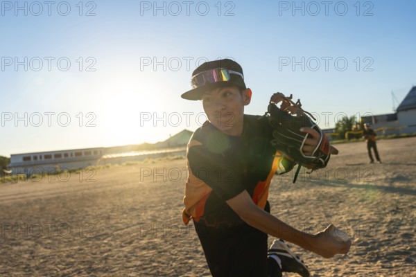 Young baseball player throwing a pitch, wearing a cap and sports goggles, with a baseball glove on his hand and a ball in motion, on a dusty field during a golden hour game