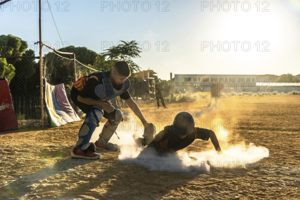 Young baseball player slides into base, sending up a cloud of dust as catcher waits on the dirt field, both backlit by warm sunset light during a youth game