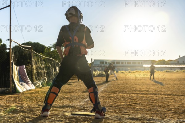 Catcher in full gear stands on a dusty baseball field at sunset, silhouetted against golden light as teammates and opponents blur in the background during evening play