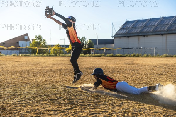 Baseball player is sliding into base creating a dust cloud while a defensive player is jumping high attempting to catch the ball during a youth game