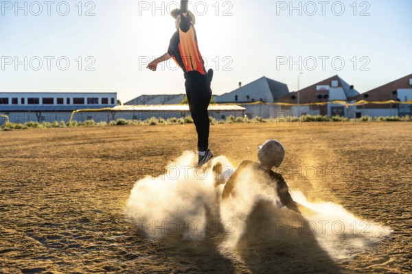 Baseball players engaging in a dynamic game, one jumping to avoid a slide while the other creates a cloud of dust, showing competitive action and teamwork on a sunny day