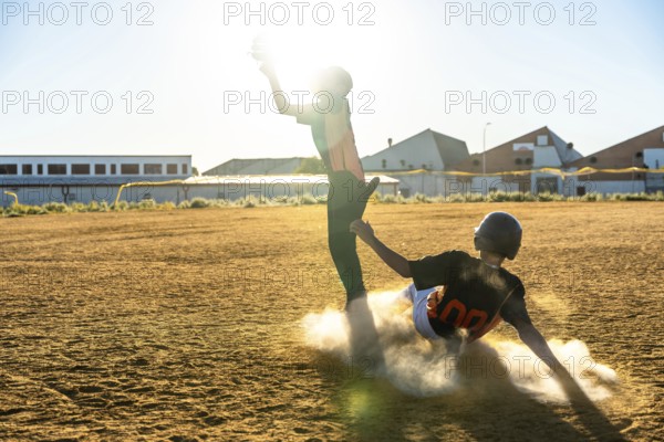 Two young baseball players are competing on a dusty field, one sliding vigorously into a base and kicking up dust while the other attempts to catch a ball, with golden sunlight in the background