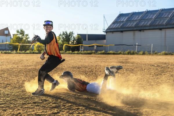 Young baseball players competing on a dusty field, one sliding head first into a base while an opponent fields the ball, engaging in competitive play and sport