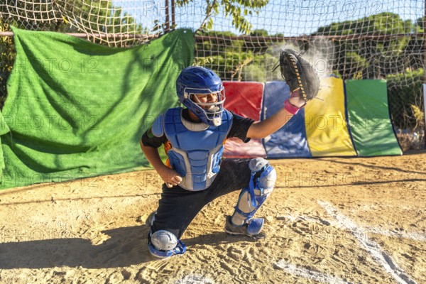 Boy catcher wearing helmet, chest protector, and leg guards, catching a baseball with glove, creating dust on an outdoor dirt field during a sports game