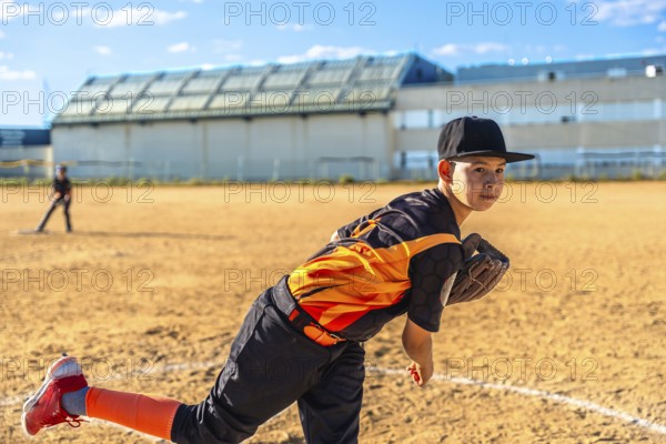 Young boy in baseball uniform and cap delivering a pitch from the mound on a sunlit dirt field, focused and determined while playing youth baseball outdoors under a clear sky