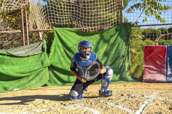 Youth baseball player wearing catcher's helmet and protective equipment, ready to catch at home plate on a sunny day at the field, representing training and competitive sport
