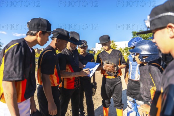 Baseball coach in black and orange uniform reviews strategy on a clipboard with attentive youth players in uniforms on the outfield under a clear blue summer sky