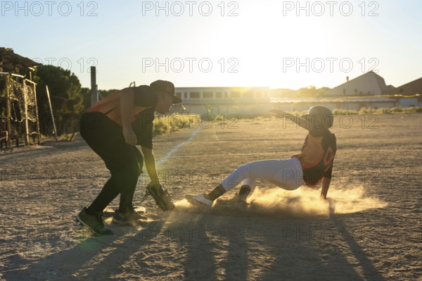 Baseball player sliding into a base, creating a cloud of dust, with another player preparing to catch the ball during a competitive game on a sunny field at sunset