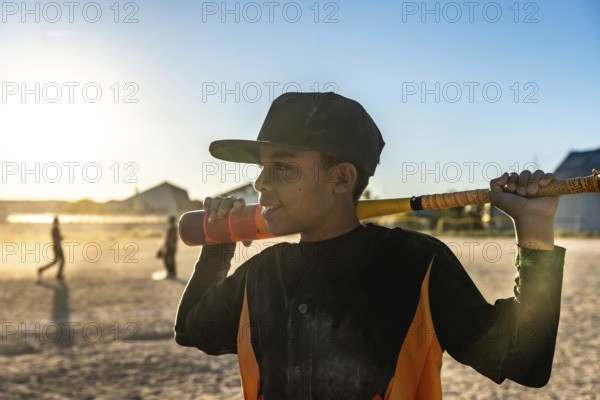 Young male baseball player holding a bat over his shoulder, wearing a cap and jersey, standing on a dusty field with other players in the background during golden hour