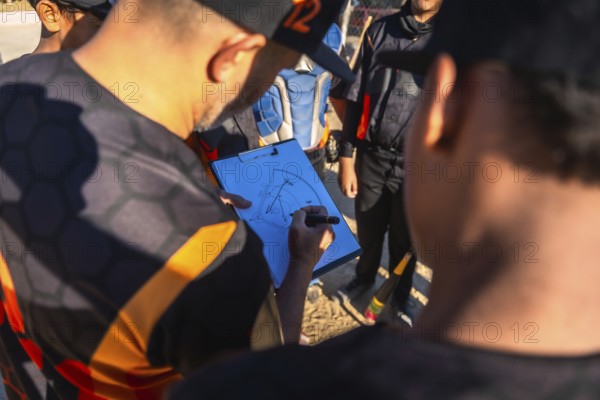 Baseball coach in uniform drawing game strategy on clipboard while explaining plays to youth team in a huddle on a sunny field, demonstrating leadership and teamwork