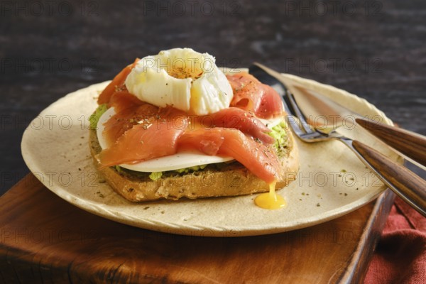 A plate holds a piece of toast topped with sliced smoked salmon and a poached egg. Yellow yolk runs down the side, and herbs are sprinkled on top. A fork and knife sit nearby on a wooden table