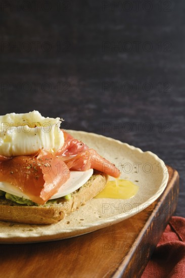 A plate holds a piece of toast topped with salmon, cream, and herbs. The setting appears casual and inviting, suggesting it is breakfast time. The wooden plate adds a rustic touch to the meal