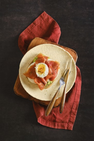 Top view of toasted bun with avocado, cheese topped with smoked salmon and poached egg. Utensils are arranged alongside the plate set on a red cloth napkin. The scene showcases a simple meal setup