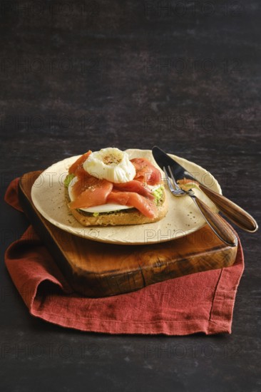 Sandwich with avocado and goat cheese topped with smoked salmon and a poached egg. The plate sits on a wooden board with a red cloth underneath. A knife and fork are placed next to the plate