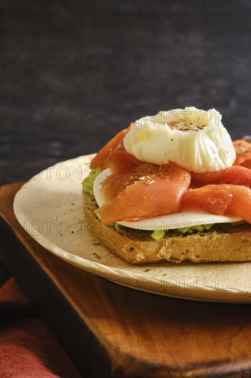 Close-up view of toasted bread with layers of avocado, smoked salmon, cheese and a poached egg on top. The dish is placed on a wooden board with dark simple background