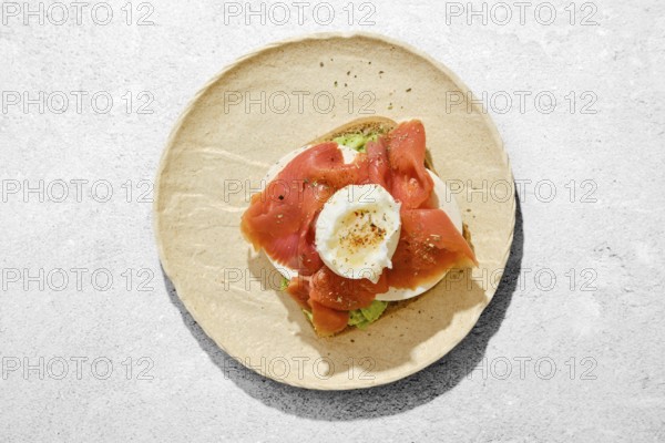 A piece of bread with avocado topped with smoked salmon, a poached egg, and seasoning on a light stone plate. This dish is prepared for breakfast and showcases fresh ingredients