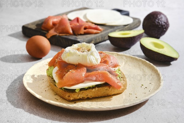 Toast topped with salmon, a poached egg, goat cheese and avocado. Sliced ingredients like salmon and cheese are on a wooden board. An egg is nearby in a kitchen setup